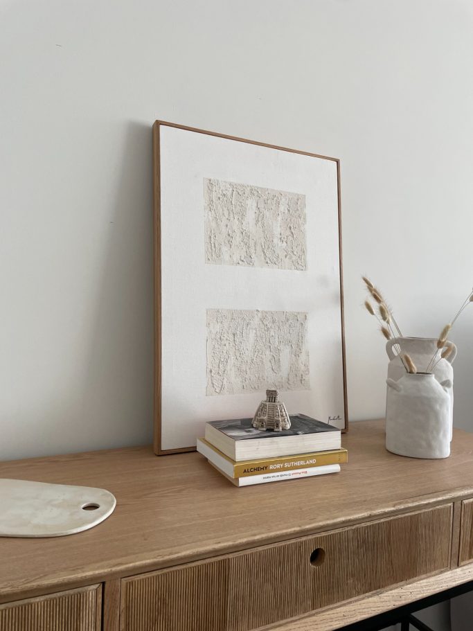 Cuadro enmarcado sobre una mesa de madera con libros y un jarrón con flores secas.